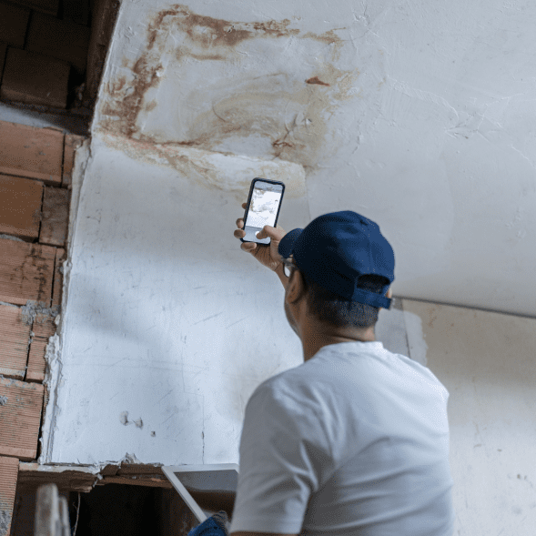 A person in a blue cap takes a photo of a water stain on a white ceiling. Exposed bricks are visible on the wall beside them.